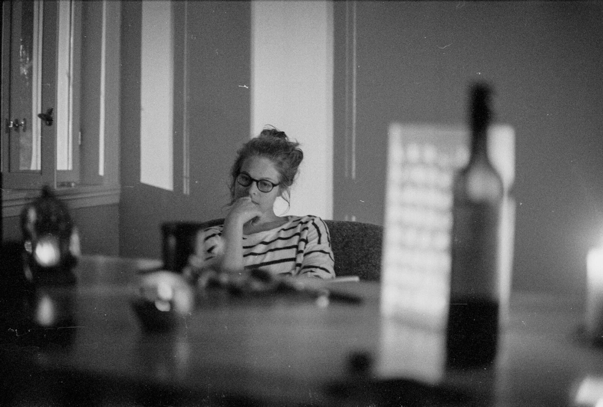 A woman is sitting on the far end of a table, engulfed in her own thoughts. There is the siluette of a bottle more close to the camera, out of focus.