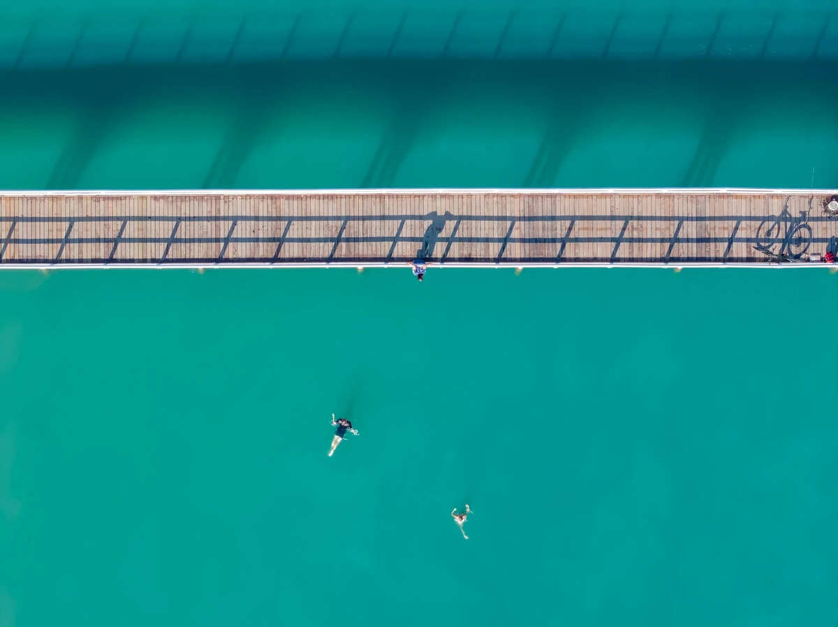A top down shot of a narrow wooden pier, sitting in aqua blue water.  Two kids are visible swimming in the water looking up at the pier, where a 3rd kid hangs off the outside of the rails, trying to build the courage to jump