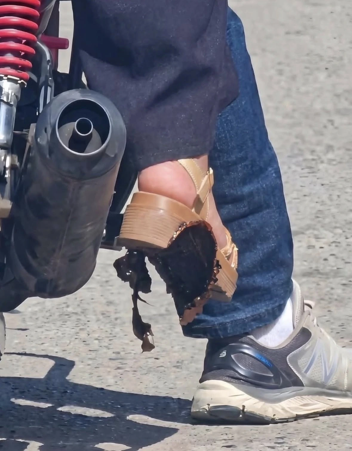 A photo of two feet, belonging to two different people sitting on the same motorbike. One has a sneaker on, the other has a platform shoe with a melted plastic sole, probably from the hot exhaust.