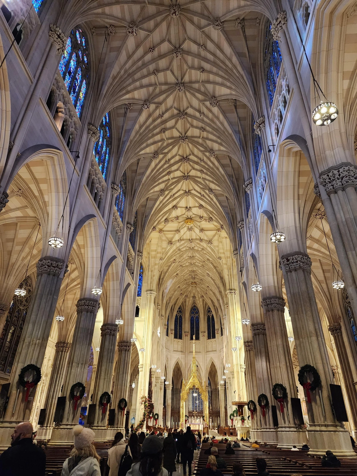 View of the altar from the entrance, showing much of the length of the ribbed vaulted ceiling and stained glass windows above the nave. in the distance is the apse with the altar under a gold canopy or altarpiece. on either side are columns dividing the main nave from the side naves, and there are people filling the aisle and pews.