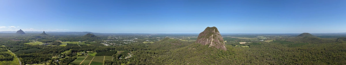 A panoramic drone photo showing the tree covered plains of the Glass House Mountains area, with the Glass House mountains themselves popping out here and there out of the landscape
